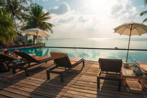 Poolside deck with lounge chairs and umbrellas next to a calm body of water, with palm trees and a bright, partly cloudy sky in the background.