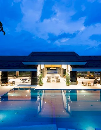 An illuminated outdoor swimming pool area with a modern building in the background, set against a dusk sky with scattered clouds.