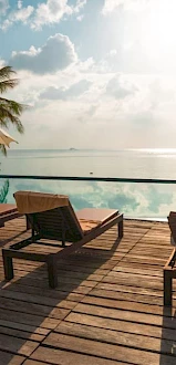 The image shows a relaxing poolside area with several lounge chairs, umbrellas, and a serene view of the sky and water reflecting the horizon.