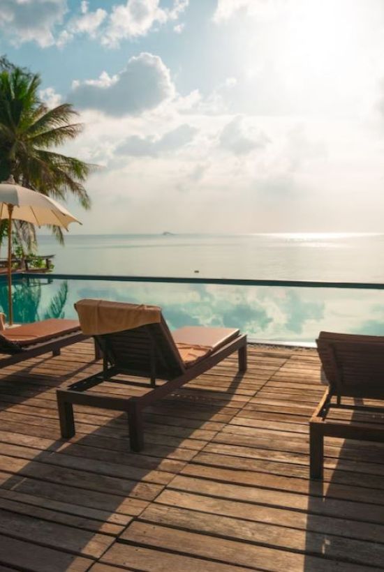 A serene tropical resort scene with sun loungers, parasols, and a swimming pool overlooking a calm sea, surrounded by lush palm trees.