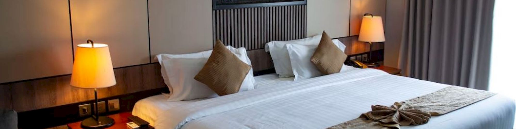 The image shows a hotel room with a large bed, two bedside lamps, and a modern wall design, lit warmly by ambient lighting.