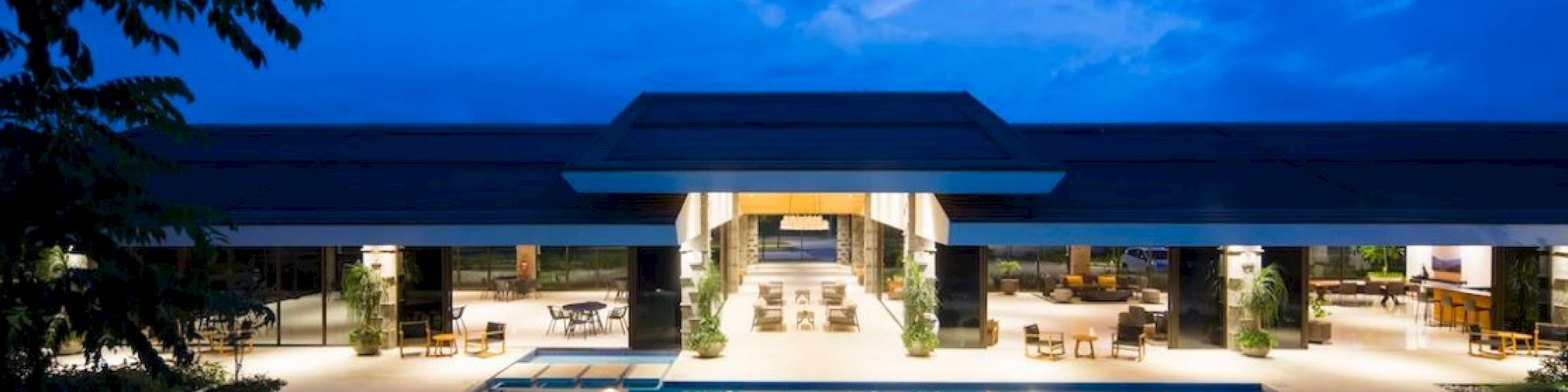 A luxurious resort with a large illuminated swimming pool and open-air common areas under a twilight sky, surrounded by modern architecture and greenery.