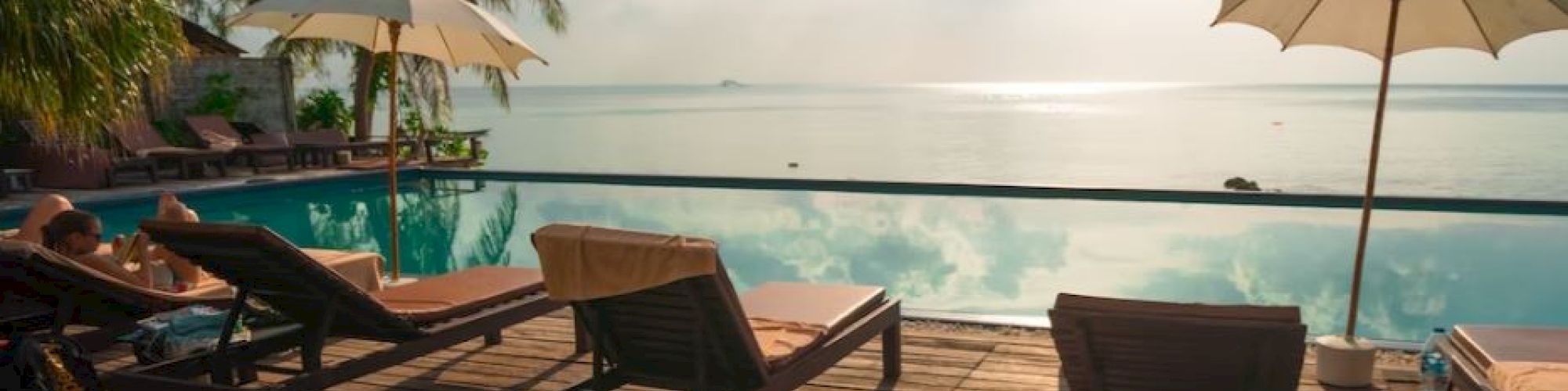 A serene poolside scene with lounge chairs, umbrellas, palm trees, and a view overlooking the ocean under a partly cloudy sky and bright sun.