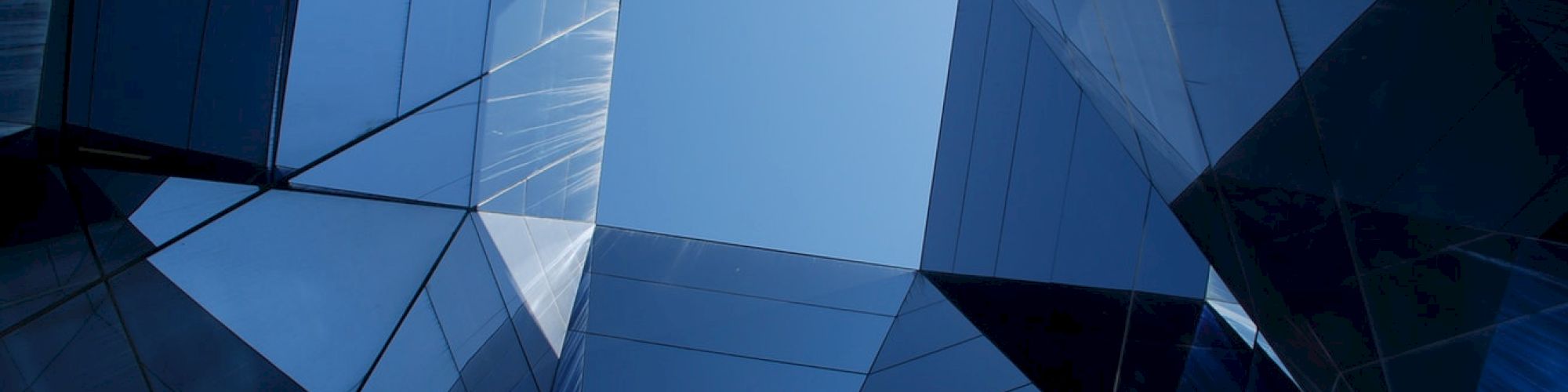 The image shows a geometric pattern formed by glass panels, reflecting and warping, creating an abstract view with a small portion of clear sky visible.