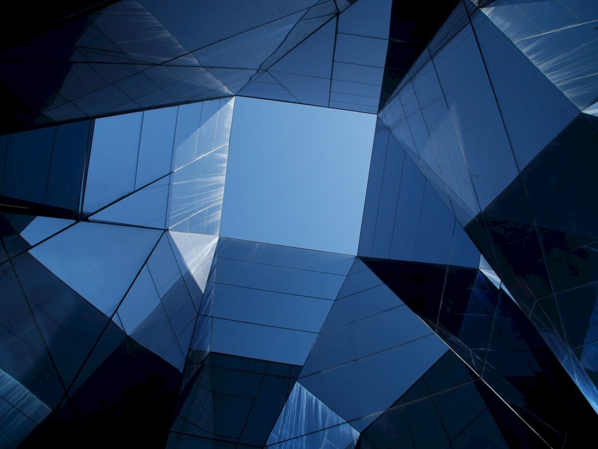 The image shows a geometric arrangement of reflective glass panels, forming an angular structure with a clear blue sky visible through the center.