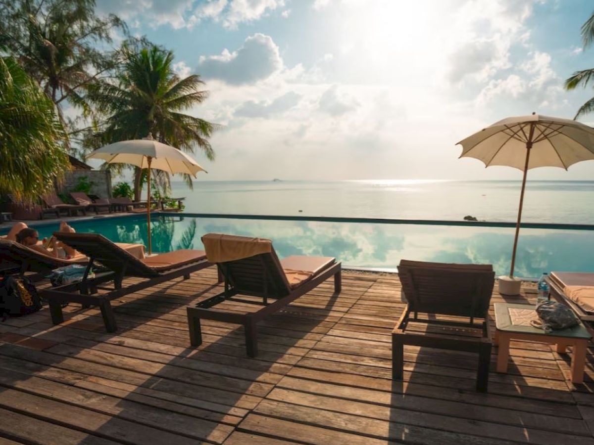 A serene poolside scene with lounge chairs and umbrellas on a wooden deck overlooking a peaceful body of water, surrounded by palm trees.