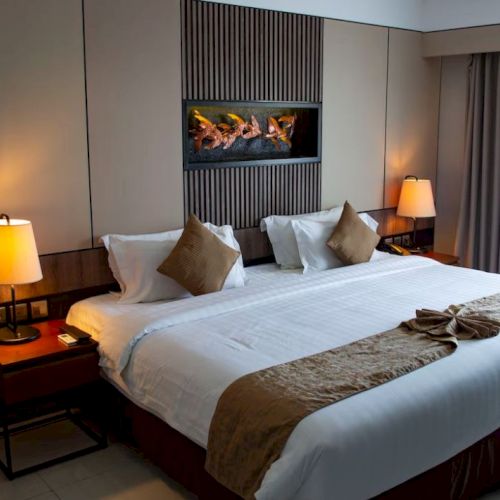 A modern hotel room features a large bed with pillows and throws, two bedside tables with lamps, and a wall-mounted fireplace.