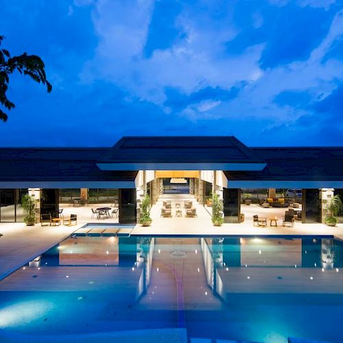 A luxurious building with large windows and an outdoor swimming pool is illuminated at twilight under a partly cloudy sky.