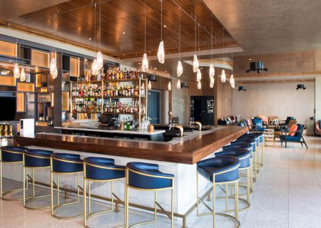 This image shows a modern bar with a wooden counter, blue barstools, a range of liquor bottles, hanging lights, and a lounge area in the background.