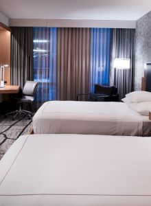 The image shows a modern hotel room with two neatly made beds, a flat-screen TV, a desk with a chair, and tasteful lighting and decor.