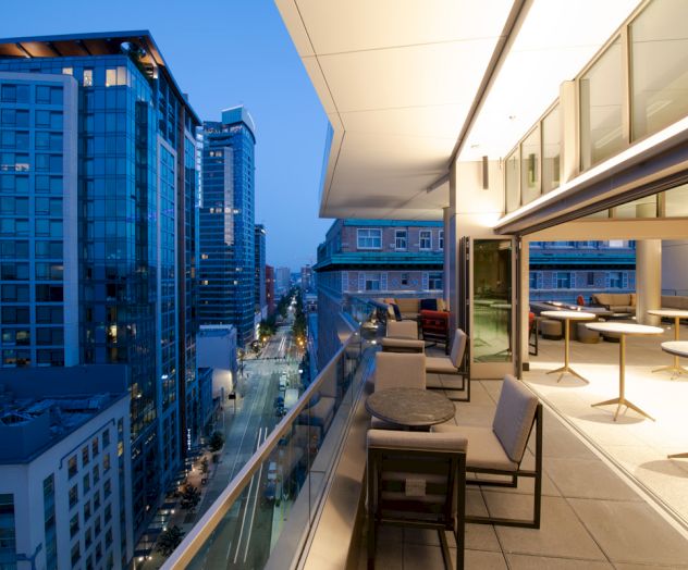 A modern balcony with tables and chairs overlooks a city street lined with tall buildings illuminated during twilight.