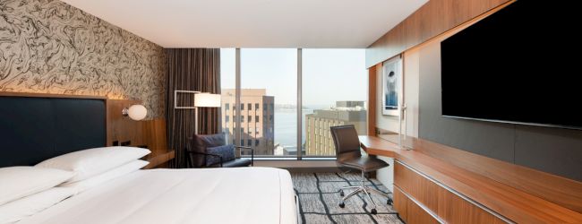 A modern hotel room with a large bed, wall-mounted TV, desk, and chair overlooking city buildings and a water view through large windows.