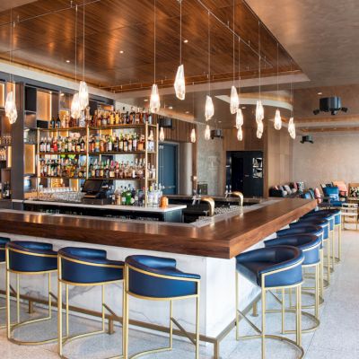 The image shows a modern bar area with a well-stocked bar, blue bar stools, hanging pendant lights, and lounge seating in the background.