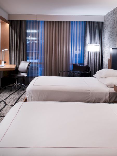 A modern hotel room with two neatly made beds, a mounted TV, a desk with a chair, and large windows with curtains in the background.