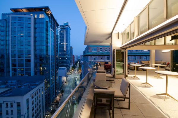 This image shows a modern cityscape with tall buildings and an outdoor terrace with tables and chairs, overlooking a street during twilight.