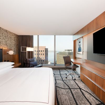 A modern hotel room features a large bed, desk with chair, and a flat-screen TV. Floor-to-ceiling window offers a cityscape and partial ocean view.