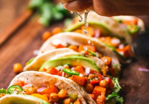The image shows a row of tacos filled with vegetables and chickpeas being topped with a spritz of fresh lime juice from a hand above.