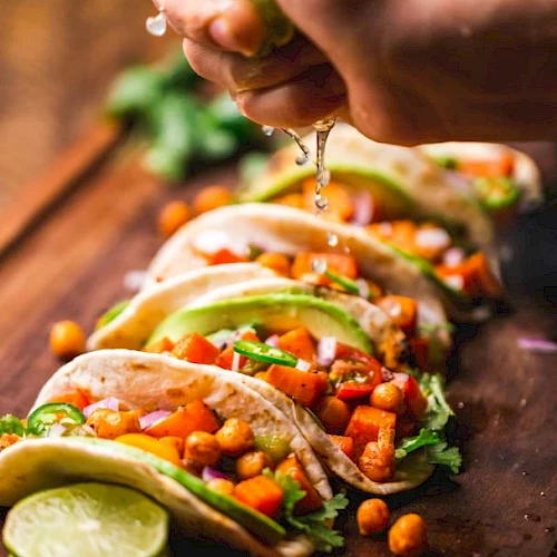 The image shows a row of tacos filled with vegetables and chickpeas being topped with a spritz of fresh lime juice from a hand above.