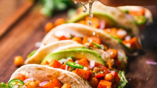 The image shows a row of tacos filled with vegetables and chickpeas being topped with a spritz of fresh lime juice from a hand above.