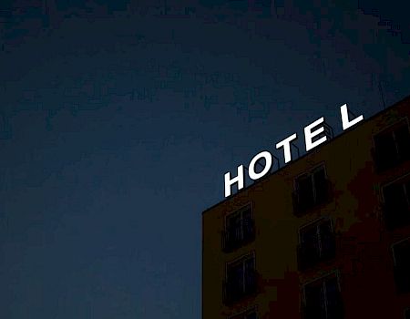 A building at dusk features a partially lit "HOTEL" sign, with the letter "T" in the hotel's sign non-illuminated.