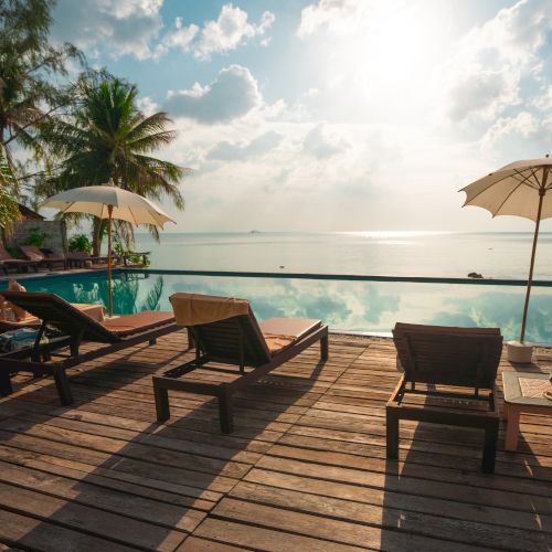 The image shows lounge chairs, umbrellas, and a wooden deck overlooking an infinity pool with a scenic ocean view and palm trees.