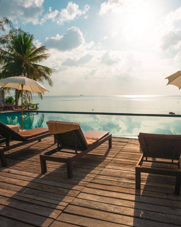 The image shows lounge chairs, umbrellas, and a wooden deck overlooking an infinity pool with a scenic ocean view and palm trees.