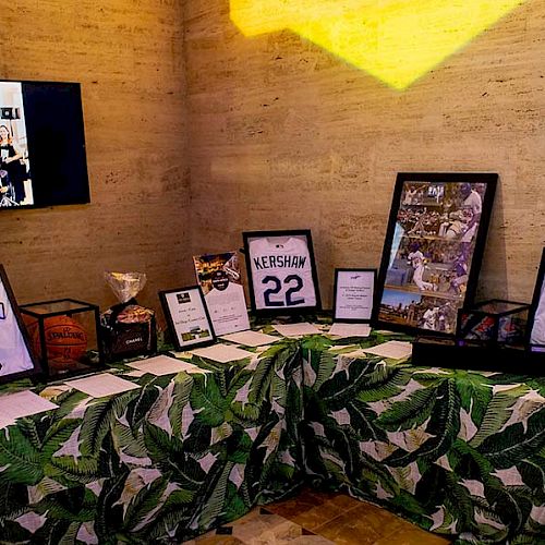 The image displays sports memorabilia on a table with a green leaf-patterned cover, including framed jerseys and certificates, plus a TV showing a photo.