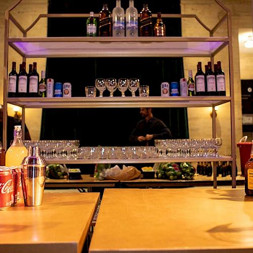 The image shows a bar setup with various bottles, cans of soda, wine bottles, glasses, and bartending equipment on shelves and a counter.