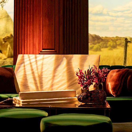 A cozy lounge area with green chairs, a table with flowers, and a large window displaying a savanna with lions in the background.