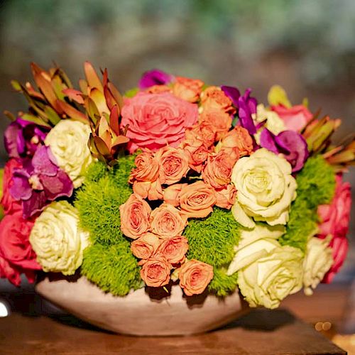 A vibrant flower arrangement in a bowl with roses, green chrysanthemums, and other colorful flowers, set against a blurred background.