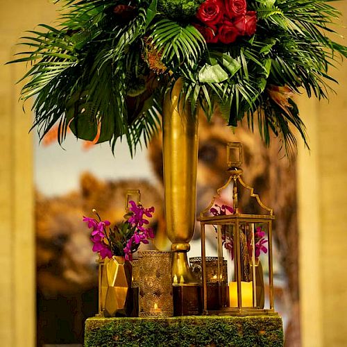 A lavish floral arrangement in a golden vase sits atop a table with a pink cloth, accompanied by smaller plants and decorative gold lanterns.