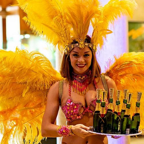 A woman in vibrant carnival attire with large yellow feathers holds a tray with several bottles of champagne, smiling for the camera.