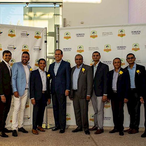 A group of ten men in formal attire stand in front of a backdrop with printed logos at an event or gathering.