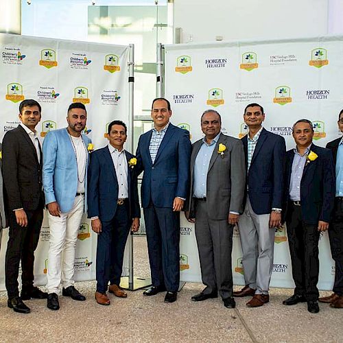 Several people, mostly men in formal attire, are posing for a group photo in front of a backdrop with logos and text, likely at an event.