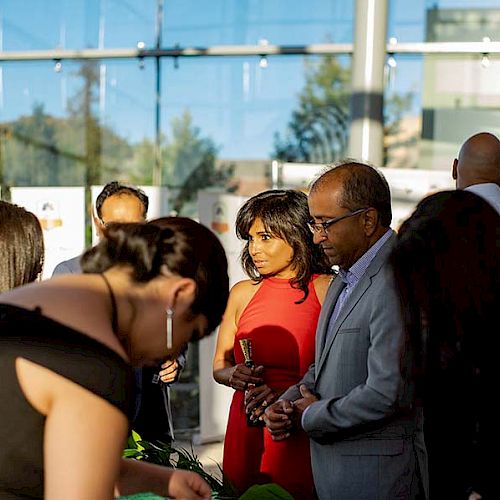 A group of people, formally dressed, socializing at an indoor event with natural light and a glass wall in the background.