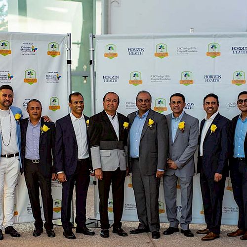 A group of people in formal attire with yellow flowers pose for a photo in front of a branded backdrop, flanked by two dancers in vibrant costumes.