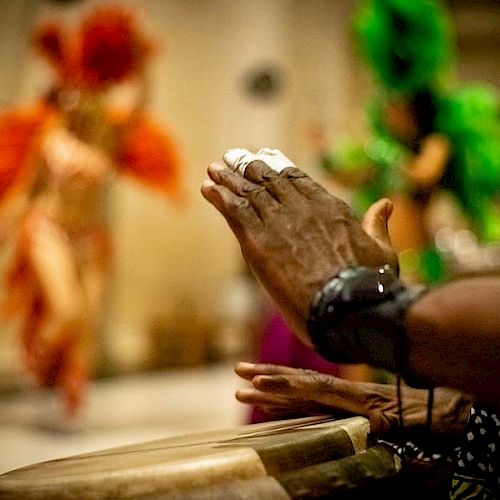 A person is playing a drum while dancers in vibrant, feathered costumes perform in the background. The scene is colorful and lively.