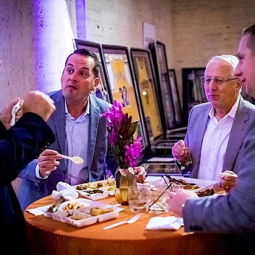 Four men are standing around a small table, eating and conversing at an event. Framed pictures can be seen in the background.