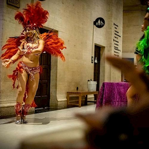 A performer in a vibrant costume with red feathers dances indoors near a doorway; another performer in green feathers is seen in the background.