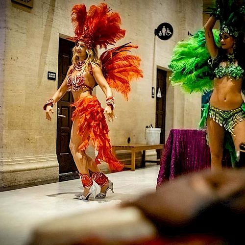 Two dancers wearing colorful feathered costumes performing indoors, one in red and the other in green, in a festive setting.