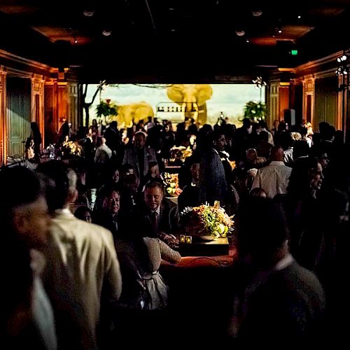 A large group of people are gathered in a dimly lit room with ornate decor, possibly for a formal event or celebration, with a colorful backdrop.