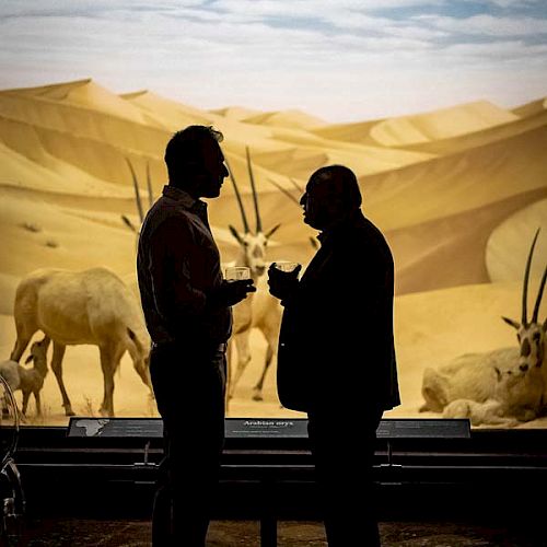 Two people are silhouetted against a desert diorama with antelope-like animals, engaging in conversation.