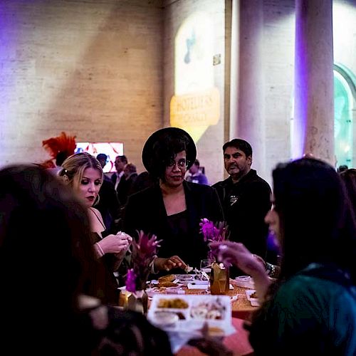 People are gathered around a table, eating and socializing in a dimly lit venue with colorful lighting and columns.