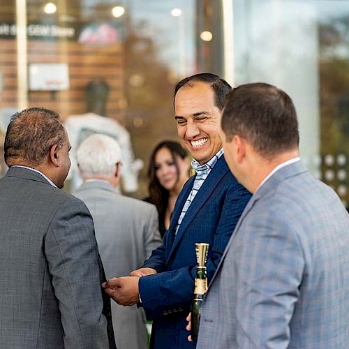 A group of people in formal attire are gathered outside, smiling and conversing near a glass storefront.