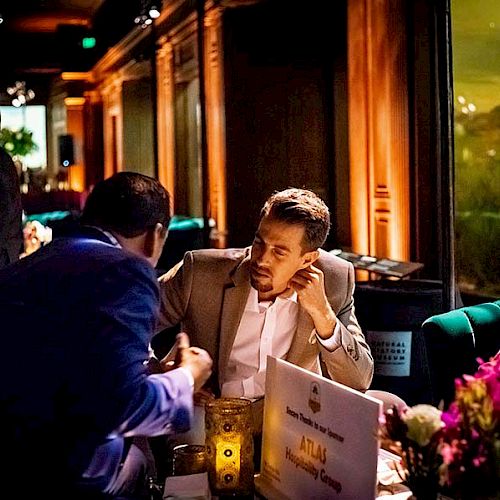 Two men are conversing at a dimly lit event or gathering, seated at a table adorned with candles and flowers.