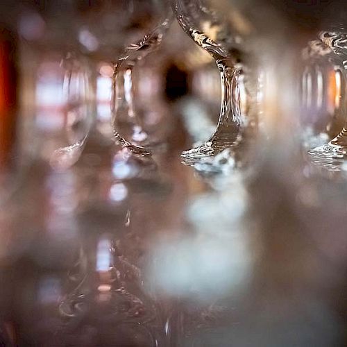 The image shows an artistic, blurred close-up of the stems of upside-down wine glasses. The focus is on light and reflections, creating an abstract effect.