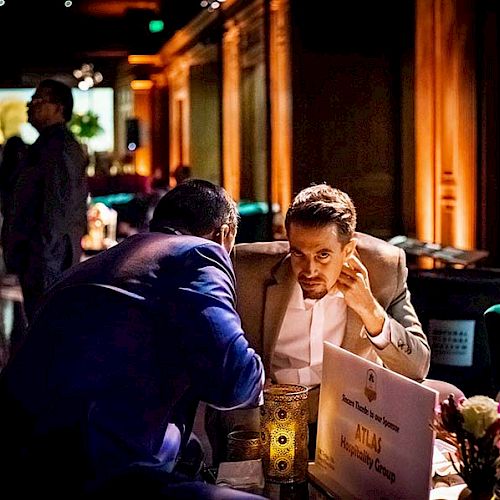 Two people are conversing closely at a dimly-lit event, with a sign and flowers on their table. The background suggests a busy establishment.