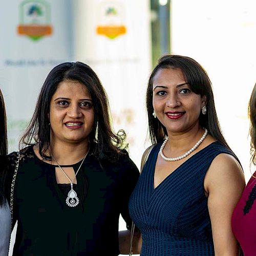 Four women are standing together, smiling at the camera, in a well-lit indoor setting. They are dressed in formal attire, suggesting an event.