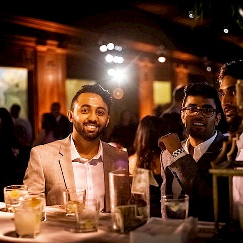 A group of men in formal attire are standing around a table with drinks, smiling and enjoying themselves in a dimly lit social setting.