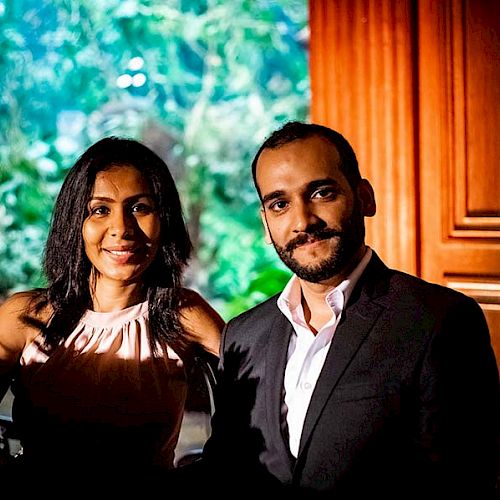 A smiling couple is posing together indoors, with a wooden panel and plants in the background.